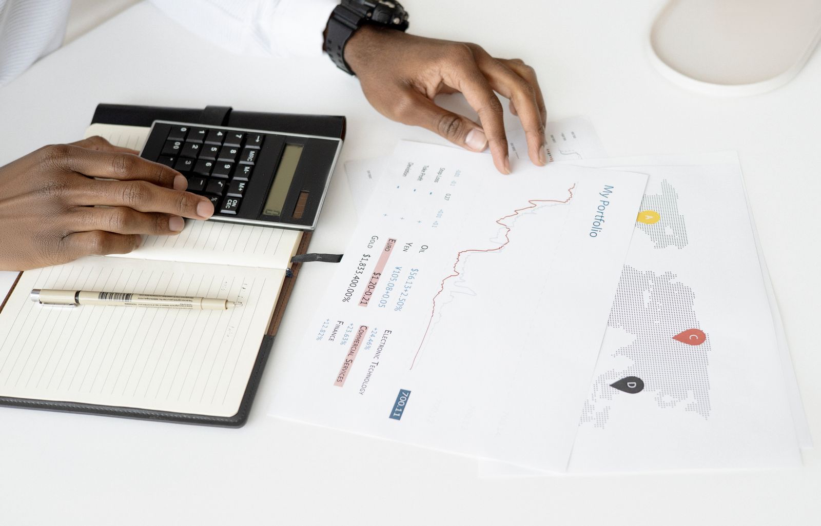 Hands using a calculator beside a notebook and printed financial charts on a desk