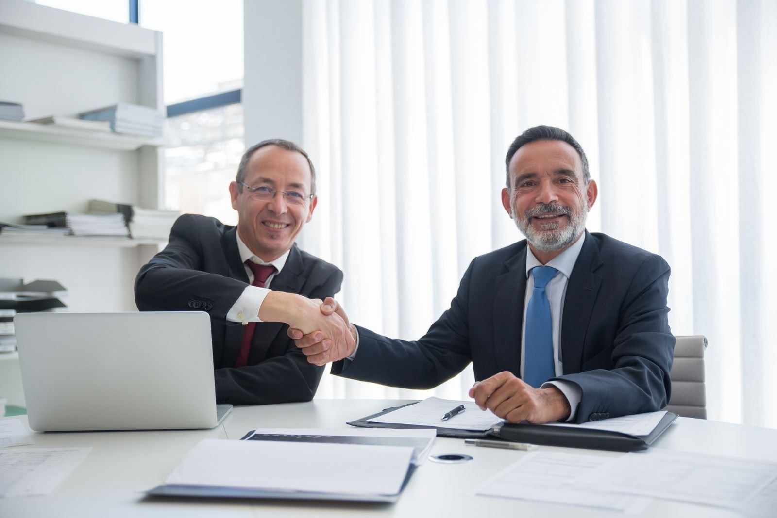 Two businessmen in suits shaking hands across a desk in a bright office